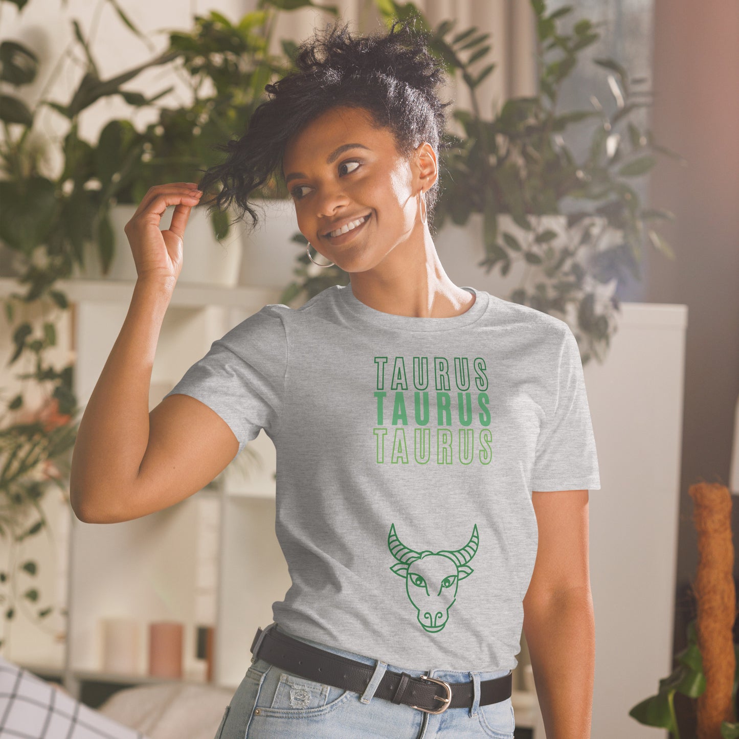 A smiling woman with curly hair, dressed in a sport grey t-shirt that reads "TAURUS TAURUS TAURUS" in green text, stands in a bright, plant-filled room. Below the text, there's a simple line drawing of a bull's head, symbolizing the Taurus zodiac sign. She looks carefree and stylish, perfectly capturing the relaxed and grounded vibe of a Taurus.