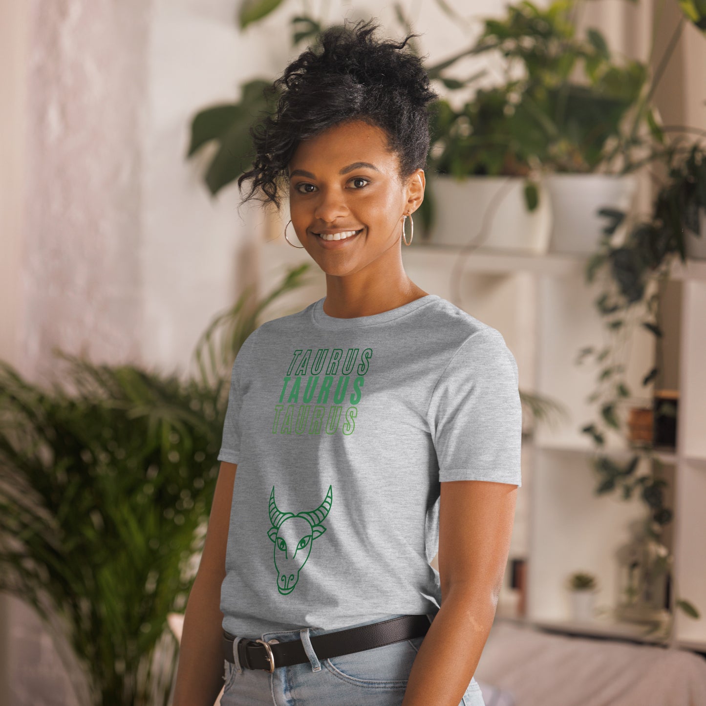 A smiling woman with curly hair, dressed in a sport grey t-shirt that reads "TAURUS TAURUS TAURUS" in green text, stands in a bright, plant-filled room. Below the text, there's a simple line drawing of a bull's head, symbolizing the Taurus zodiac sign. She looks carefree and stylish, perfectly capturing the relaxed and grounded vibe of a Taurus.
