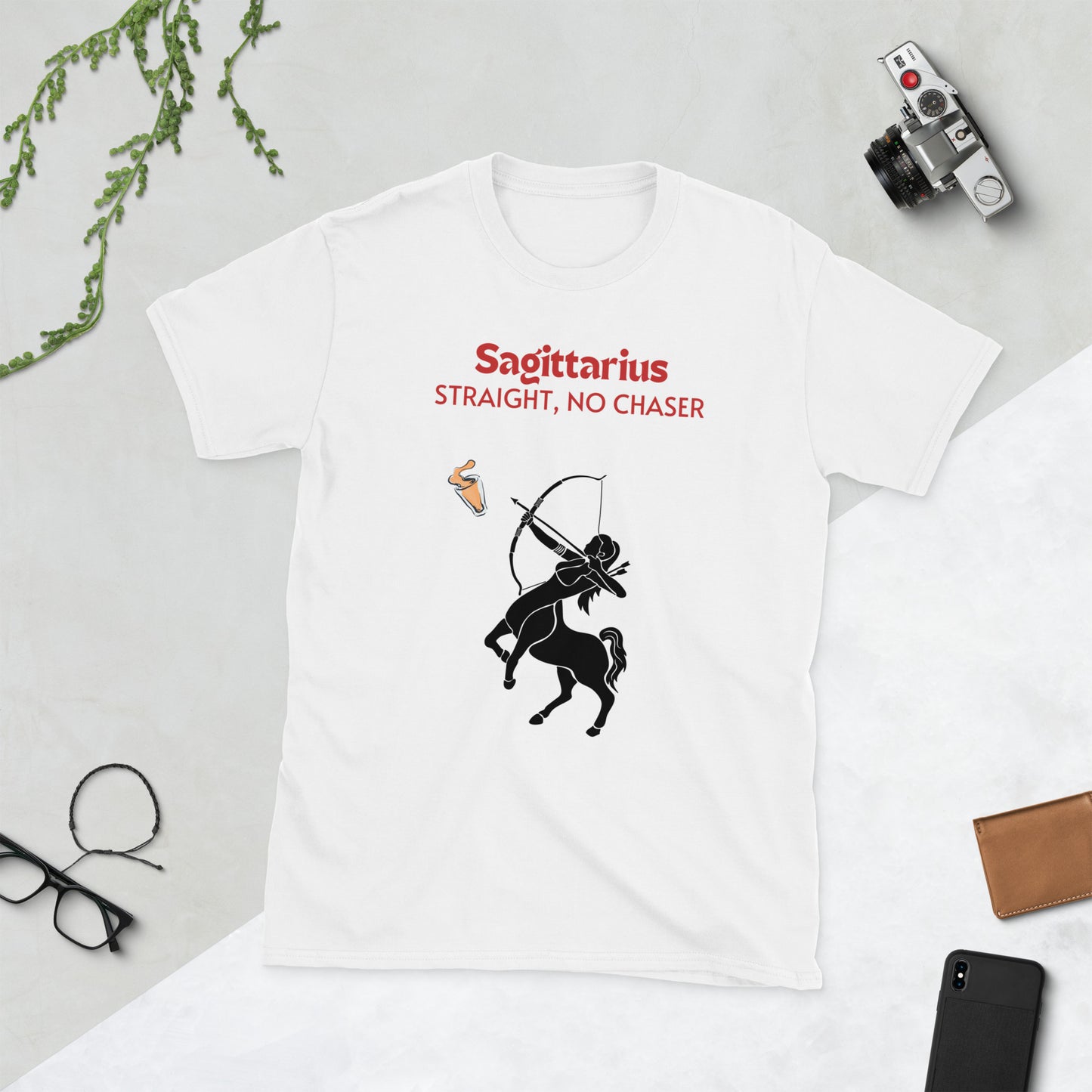 A white t-shirt lying flat on a light background, featuring a bold "Sagittarius - Straight, No Chaser" design. The graphic showcases a black centaur archer aiming at a drink, embodying the fearless and straightforward nature of Sagittarius. The shirt is surrounded by stylish accessories, including a camera, sunglasses, and a wallet.