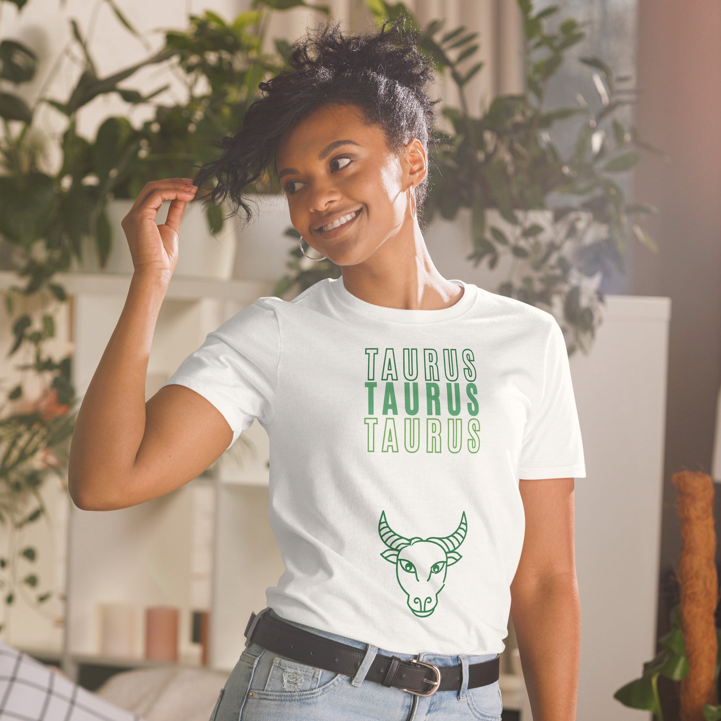 A smiling woman with curly hair, dressed in a white t-shirt that reads "TAURUS TAURUS TAURUS" in green text, stands in a bright, plant-filled room. Below the text, there's a simple line drawing of a bull's head, symbolizing the Taurus zodiac sign. She looks carefree and stylish, perfectly capturing the relaxed and grounded vibe of a Taurus.
