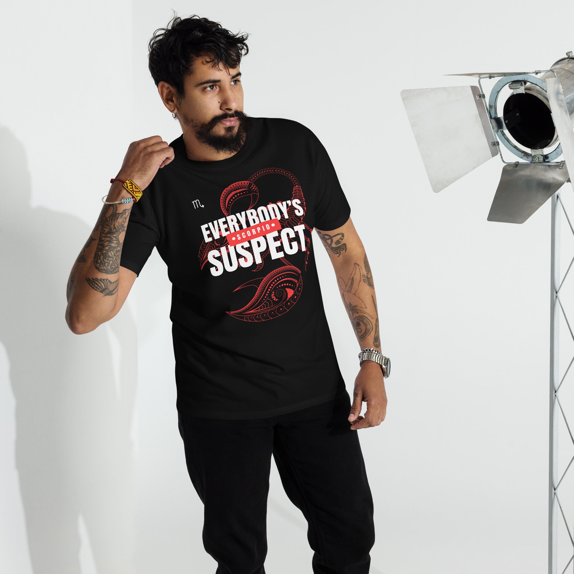 A man wearing a black t-shirt with the phrase "Everybody's Suspect" in bold red and white letters, and a detailed scorpion design in the background, indicating it is a Scorpio-themed shirt. He is standing in a casual pose with one hand raised, wearing black jeans, and accessorized with bracelets and a watch.