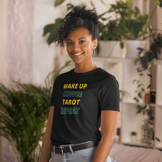 A woman wearing a black t-shirt with the text "Wake Up, Coffee, Tarot, Repeat" in colorful letters. She is standing in a cozy, plant-filled room, smiling, with her hair up and wearing hoop earrings, a belt, and jeans.